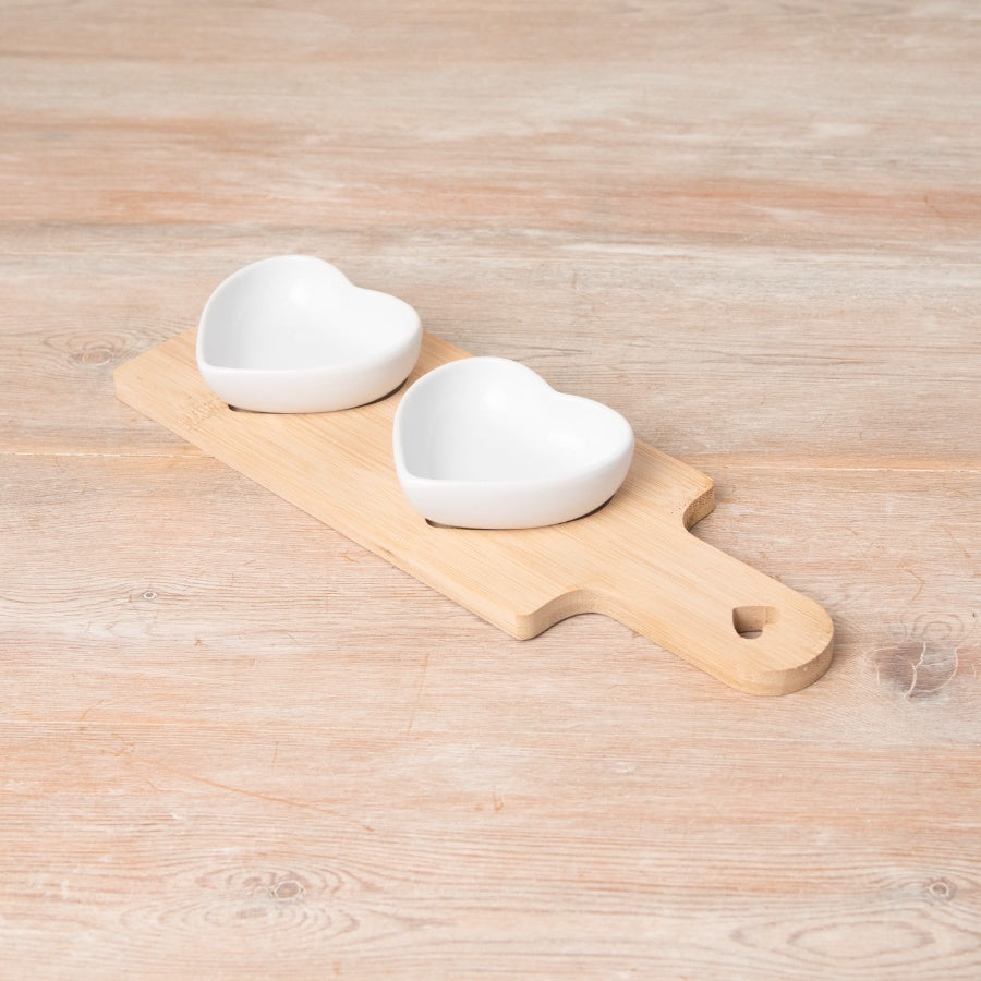 Two white ceramic heart-shaped bowls sitting on a natural wooden serving board with heart cut-out handle