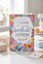 Cousin birthday card with bright colourful floral border and silver foil lettering reading For You Cousin Wishing You the Loveliest Birthday Ever, styled on a table with gift boxes and confetti