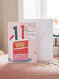 11th birthday card with illustrated cake slice, leopard print candles and gold stars on pink background with white envelope