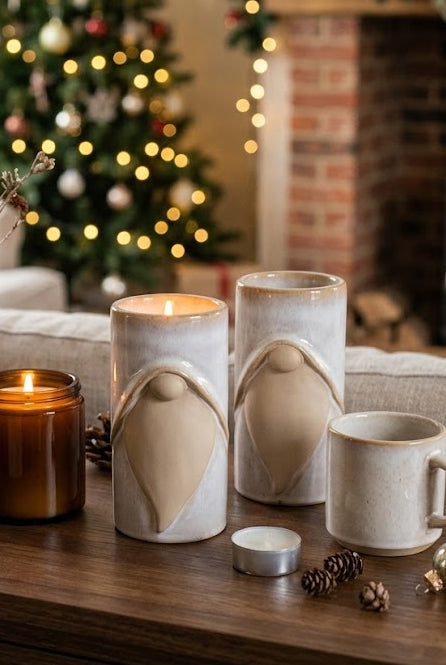 A cozy holiday living room scene with a wooden table holding ceramic gnome-shaped candle holders, a lit amber candle, pinecones, and dried stems in a vase, with a decorated Christmas tree and fireplace in the background.