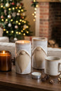 A cozy holiday living room scene with a wooden table holding ceramic gnome-shaped candle holders, a lit amber candle, pinecones, and dried stems in a vase, with a decorated Christmas tree and fireplace in the background.