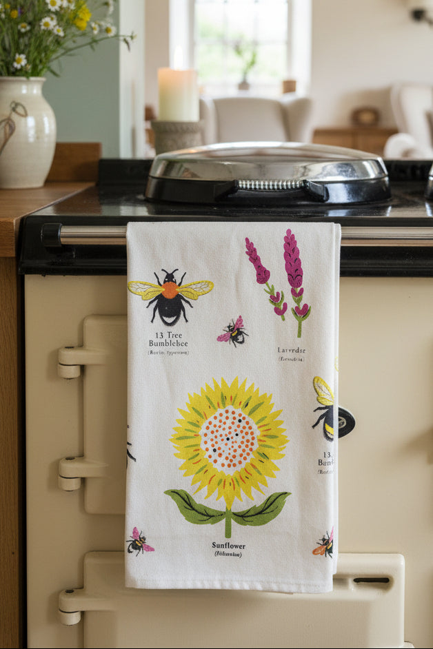 A cotton tea towel with a Garden Bees pattern, hanging on a wooden stand in a home setting. The towel features a large yellow sunflower, a bumblebee, and lavender flowers on a cream background, with the product packaging card visible at the top.