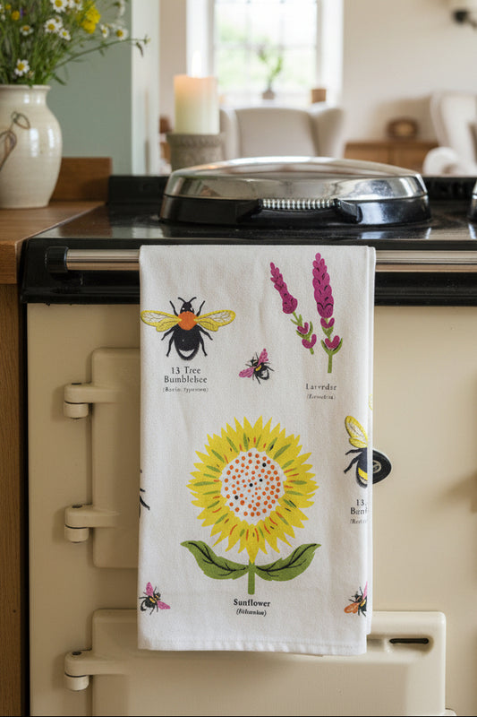 A cotton tea towel with a Garden Bees pattern, hanging on a wooden stand in a home setting. The towel features a large yellow sunflower, a bumblebee, and lavender flowers on a cream background, with the product packaging card visible at the top.