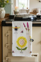 A cotton tea towel with a Garden Bees pattern, hanging on a wooden stand in a home setting. The towel features a large yellow sunflower, a bumblebee, and lavender flowers on a cream background, with the product packaging card visible at the top.