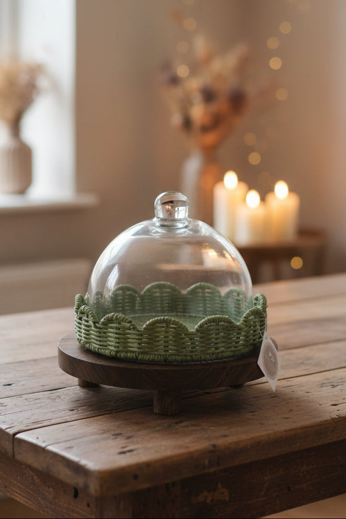 A green woven basket-style cloche with a glass dome lid, displayed on a round wooden stand. A white hanging tag is attached to the front, and the product is photographed on a dark tabletop with a laptop and shelf visible in the background.