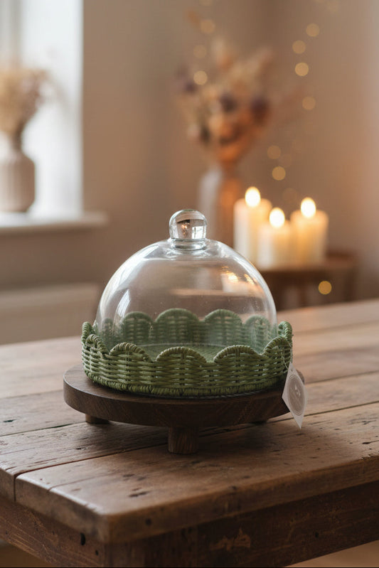 A green woven basket-style cloche with a glass dome lid, displayed on a round wooden stand. A white hanging tag is attached to the front, and the product is photographed on a dark tabletop with a laptop and shelf visible in the background.