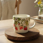 A ceramic mug with a hand-painted strawberry and white flower pattern, displayed on a round wooden riser.