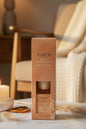 A tall rectangular box for an Eden Oud & Sandalwood Reed Diffuser stands on a round wooden pedestal. The packaging is a warm peach-tan color with black text and a small clear window showing a beige cylindrical diffuser inside.