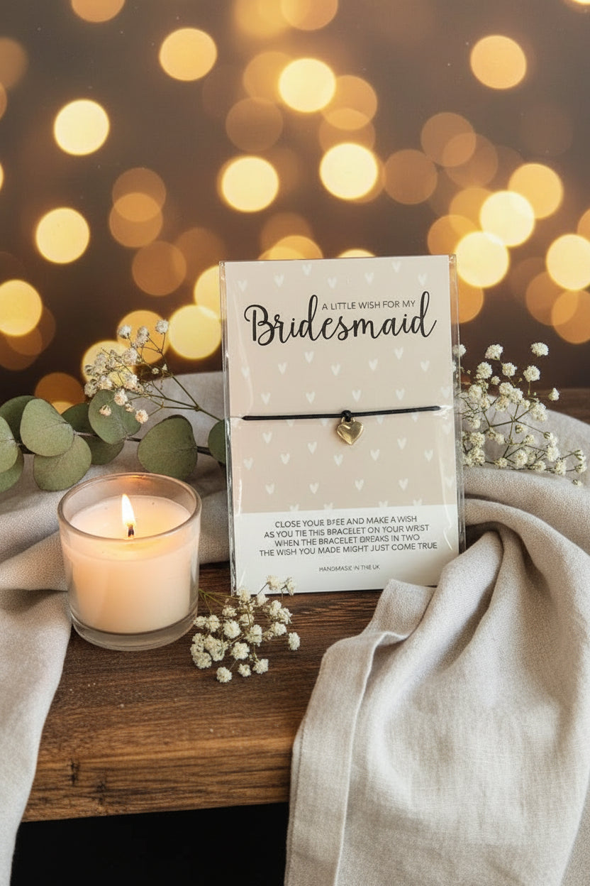 A bridesmaid bracelet displayed on a printed card inside a clear acrylic stand on a wooden table. The bracelet is a thin black cord with a small silver heart charm, and the card reads 'A Little Wish for My Bridesmaid' with additional text below.