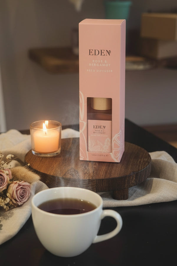 A pink Eden Rose & Bergamot reed diffuser box with a clear window showing the bottle inside, displayed on a round wooden stand on a dark table.
