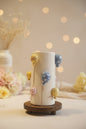A handmade ceramic cylinder candle holder with a beige body, decorated with colorful hand-painted balloons in yellow, blue, pink, and purple, sitting on a round wooden base on a dark table.