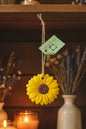 A glossy yellow flower-shaped ornament with a brown textured center and a natural jute hanging loop, shown on a round wooden surface with a small green tag attached.