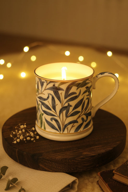A white ceramic mug with candle that fits inside with a blue botanical pattern and gold trim, sitting on a round wooden coaster on a dark table in an indoor setting.