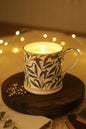 A white ceramic mug with candle that fits inside with a blue botanical pattern and gold trim, sitting on a round wooden coaster on a dark table in an indoor setting.