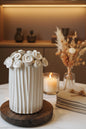 A white ceramic column-style candle holder with decorative rosettes around the rim, displayed on a round wooden base on a dark tabletop in an indoor room setting.