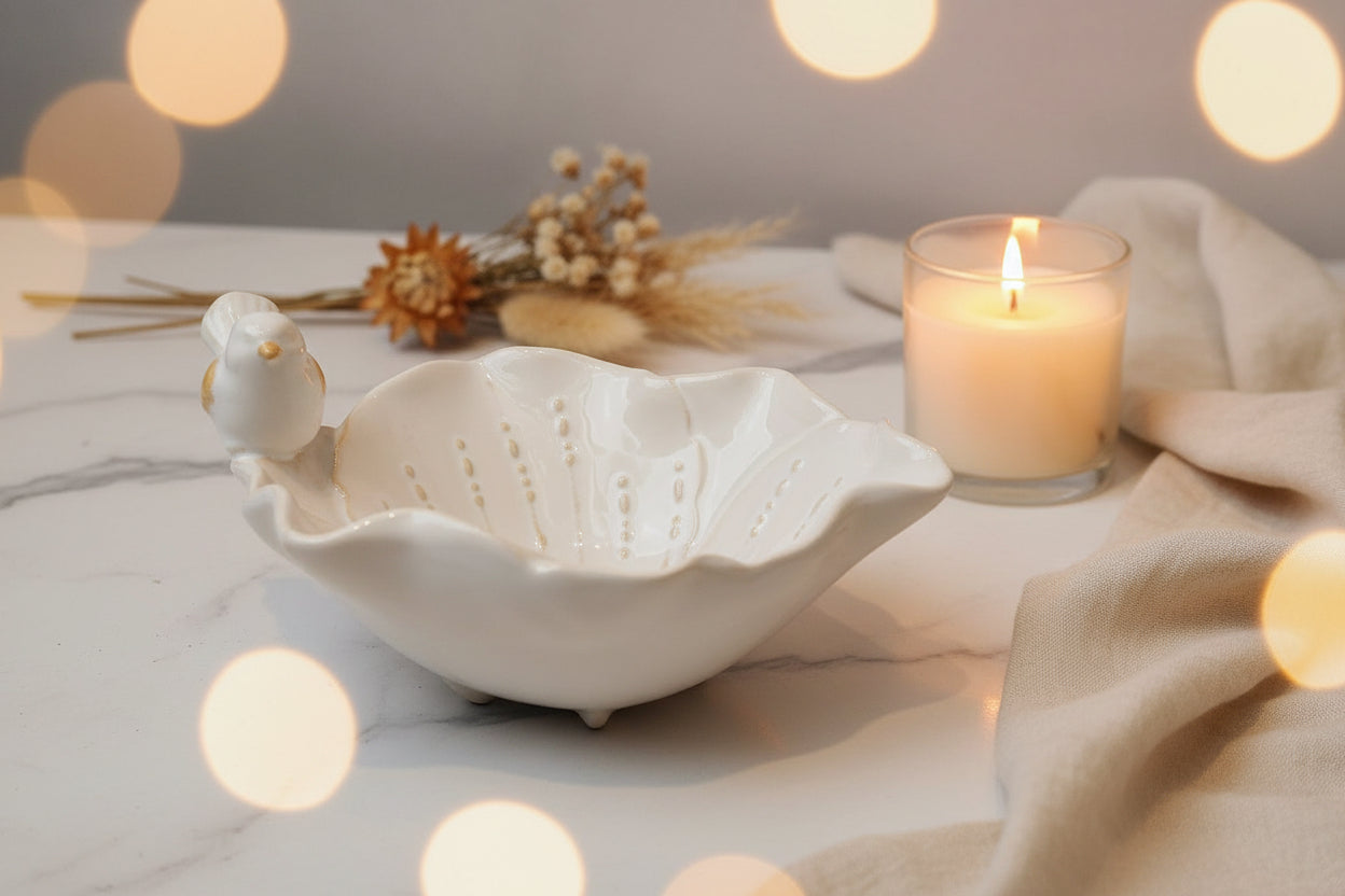 A white ceramic bird-shaped bowl with a leaf-like rim, featuring small perforated details inside and a small bird figure perched on the left side, shown on a dark wooden surface.