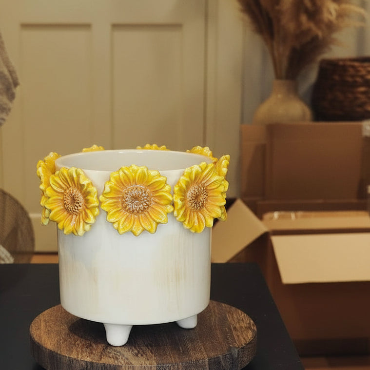 A white ceramic footed planter decorated with three large yellow sunflower petals around the rim, displayed on a round wooden coaster.