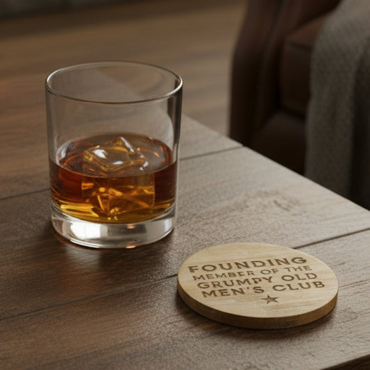 A round wooden coaster engraved with the text 'FOUNDING MEMBER OF THE GRUMPY OLD MEN'S CLUB' and a small star, shown on a dark surface.