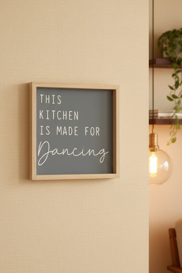 A square framed sign with a light wood frame and a frosted gray background, featuring the text 'THIS KITCHEN IS MADE FOR Dancing' in white lettering, displayed on a dark tabletop.