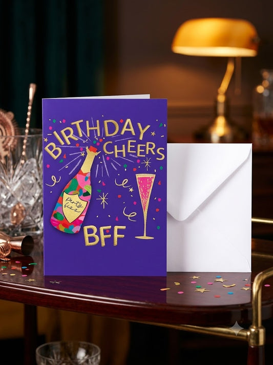 Birthday Cheers BFF card with gold foil lettering, champagne bottle and pink fizz glass on purple background with white envelope