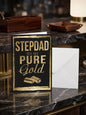Stepdad You Are Pure Gold birthday card — bold black and gold foil design, dark marble lifestyle photo with leather tray and lamp