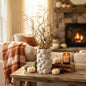 A textured white ceramic vase filled with dried brown branches sits on a rustic wooden table in a cozy living room setting with a fireplace, pumpkins, and a candle.