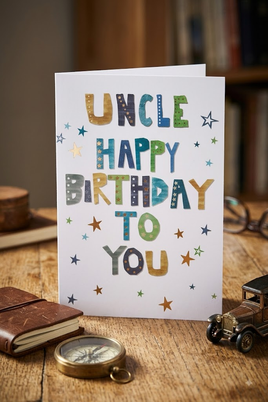 Uncle Happy Birthday To You card with bold multicoloured lettering and scattered stars on white, styled on a rustic wooden table with a compass, vintage toy car and leather notebook