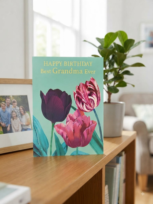 Happy Birthday Best Grandma Ever card featuring three illustrated tulips in plum, striped pink and hot pink on a teal background with gold foil lettering, displayed on a wooden sideboard beside a family photo frame and a potted plant