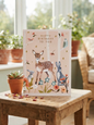 Happy Birthday To You card featuring an illustrated fawn surrounded by a mouse, blue hare and barn owl among pink, orange and teal wildflowers on a blush background with gold star details, displayed on a rustic wooden table beside a terracotta succulent pot in a bright conservatory