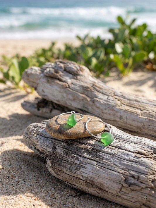 Green sea glass hoop earrings on driftwood and pebble on a sandy beach by Laura Loos