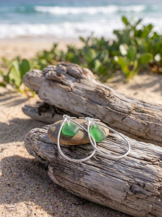 Green sea glass teardrop sterling silver earrings on driftwood and pebble on a sandy beach by Laura Loos