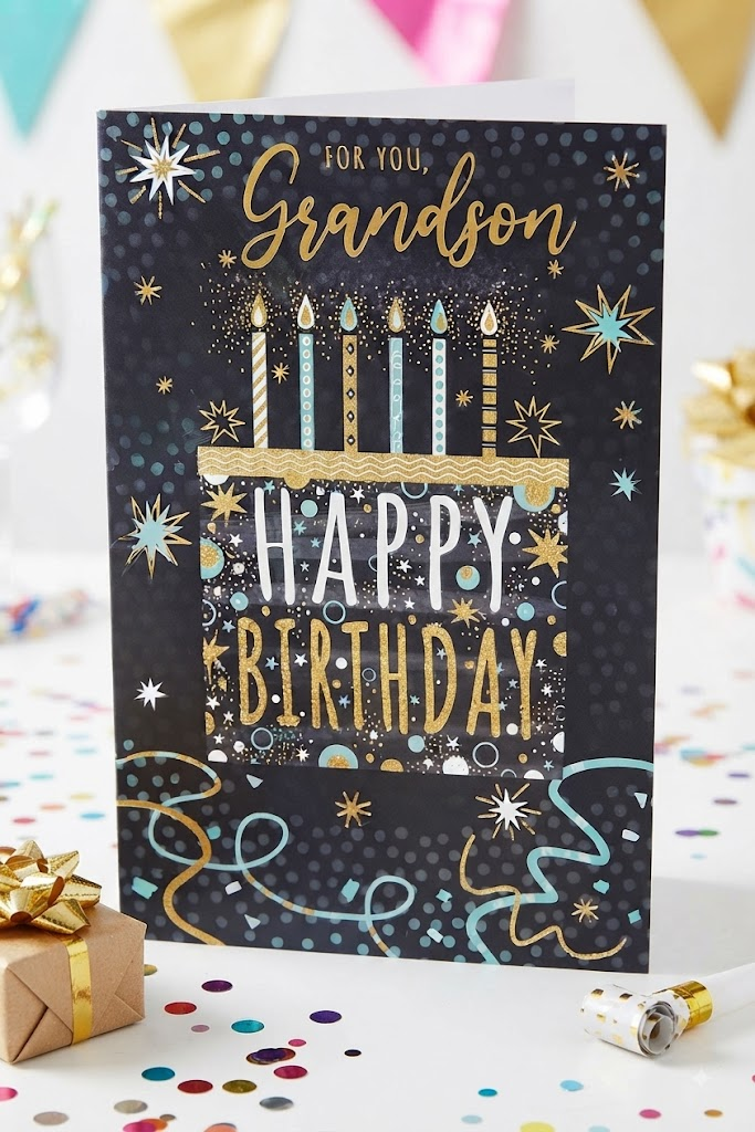 For You Grandson Happy Birthday card featuring a birthday cake with candles, gold foil star bursts and teal and gold confetti on a dark background, displayed on a white table with confetti, a gold gift box and party popper