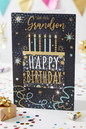 For You Grandson Happy Birthday card featuring a birthday cake with candles, gold foil star bursts and teal and gold confetti on a dark background, displayed on a white table with confetti, a gold gift box and party popper