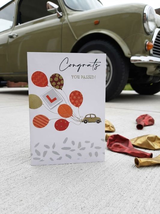 Congrats You Passed driving test card featuring an illustrated car floating with colourful patterned balloons and an L-plate, displayed on a driveway in front of a vintage olive green Mini