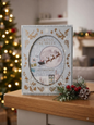 Across The Miles At Christmas card with Santa's sleigh over a full moon and gold foil botanical border on grey-blue background