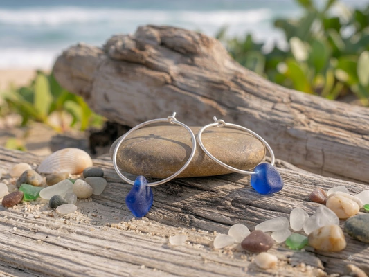 Laura Loos handmade cobalt blue sea glass drop hoop earrings in 925 sterling silver, displayed on driftwood with sea glass pebbles on a sandy beach
