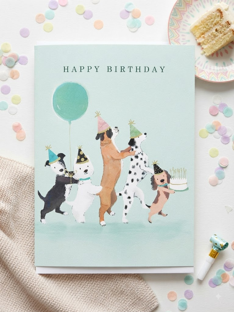 Happy Birthday card featuring four illustrated dogs in party hats dancing with a teal balloon and birthday cake on a mint green background, displayed flat on a white surface surrounded by pastel confetti circles beside a slice of cake and a party blower