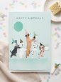 Happy Birthday card featuring four illustrated dogs in party hats dancing with a teal balloon and birthday cake on a mint green background, displayed flat on a white surface surrounded by pastel confetti circles beside a slice of cake and a party blower