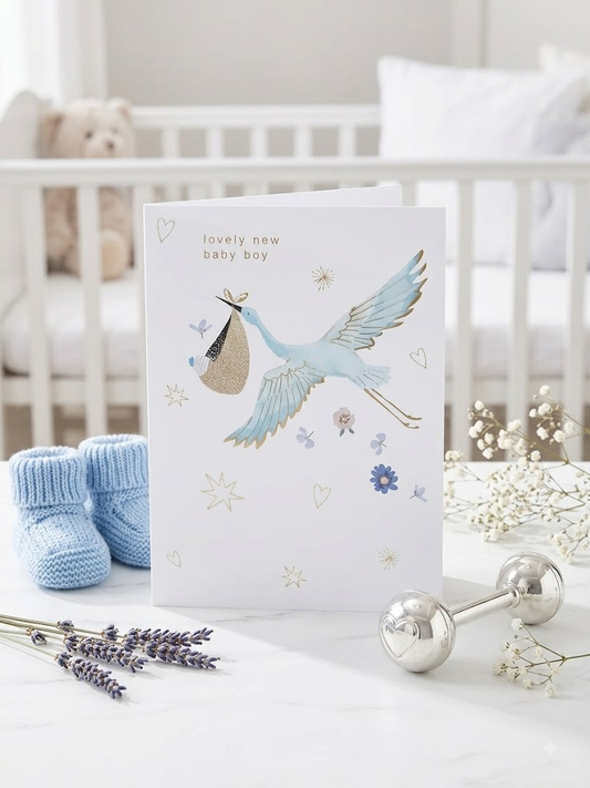 Lovely New Baby Boy greeting card with illustrated blue stork carrying a bundle, gold star and floral details, displayed in a nursery setting with blue baby booties and a silver rattle