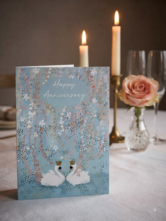 Happy Anniversary card featuring two elegant crowned swans on a teal blue background with cascading cherry blossom and floral details, displayed on a candlelit dinner table with a pink rose and champagne glasses