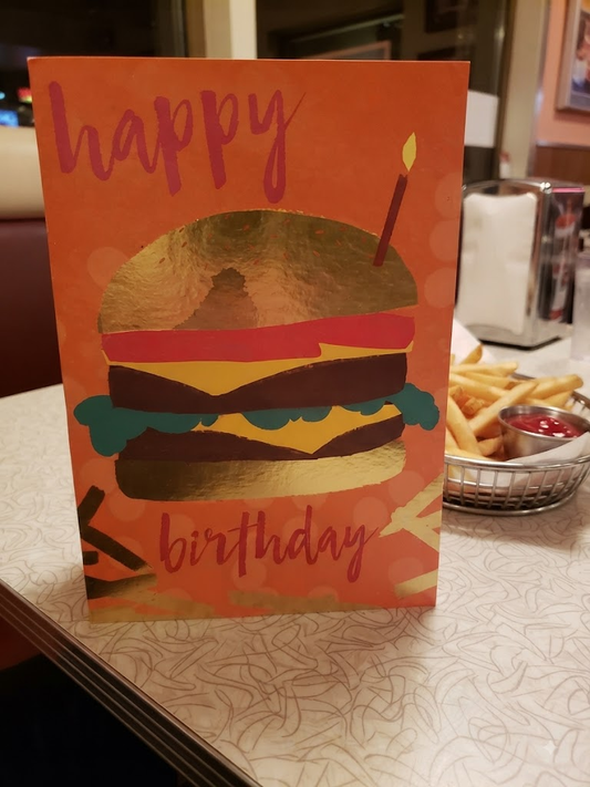 Happy Birthday burger greeting card featuring a bold illustrated cheeseburger with a birthday candle and gold foil details on a vibrant orange background, displayed on a diner table with chips and ketchup in the background