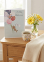 As You Recover From Your Operation card featuring an illustrated teddy bear floating with patterned balloons in dusky pink, mauve and navy with a 'with love' tag, displayed on a bedside table with a floral mug of tea, yellow gerberas and chamomile flowers