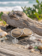 Laura Loos handmade frosted white sea glass drop hoop earrings in 925 sterling silver, displayed on driftwood with sea glass pebbles on a sandy beach