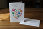 Rainbow paw print heart pet sympathy card standing upright spine-left slightly open in a cosy lifestyle setting