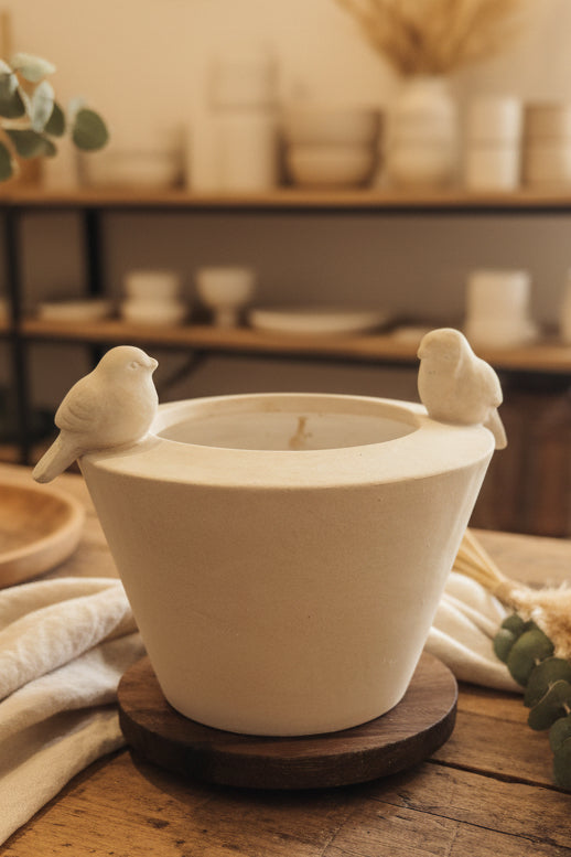A cream-colored ceramic candle holder shaped like a planter with two small bird figurines perched on the rim, displayed on a wooden coaster on a rustic wooden table.