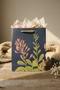 A navy blue gift bag with a botanical illustration of pink flowers and green leaves, standing on a rustic wooden surface with a beige woven blanket and dried flowers in the background.