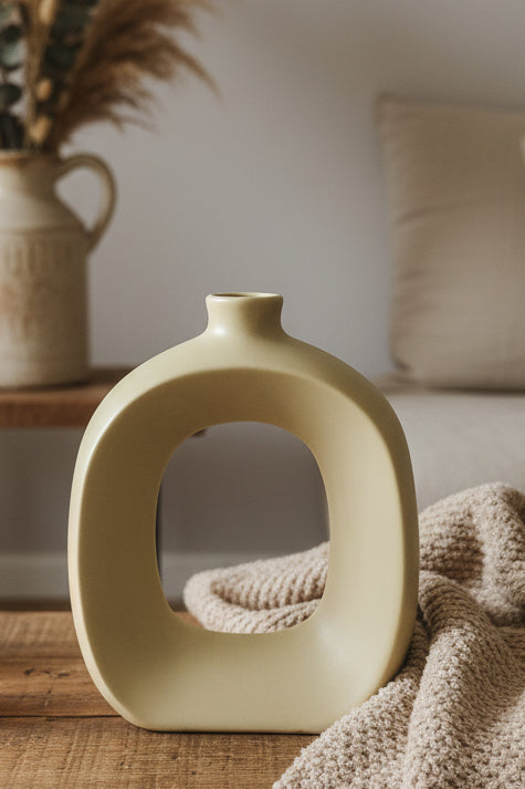 A matte cream-colored ceramic vase with a unique rounded square shape and a central cutout, styled on a wooden table next to a chunky knit blanket.