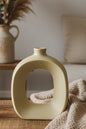 A matte cream-colored ceramic vase with a unique rounded square shape and a central cutout, styled on a wooden table next to a chunky knit blanket.
