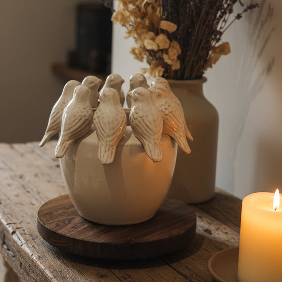 A ceramic bowl with six sculpted birds perched on top, displayed on a round wooden tray on a rustic table next to a lit candle, with a vase of dried flowers in the background.